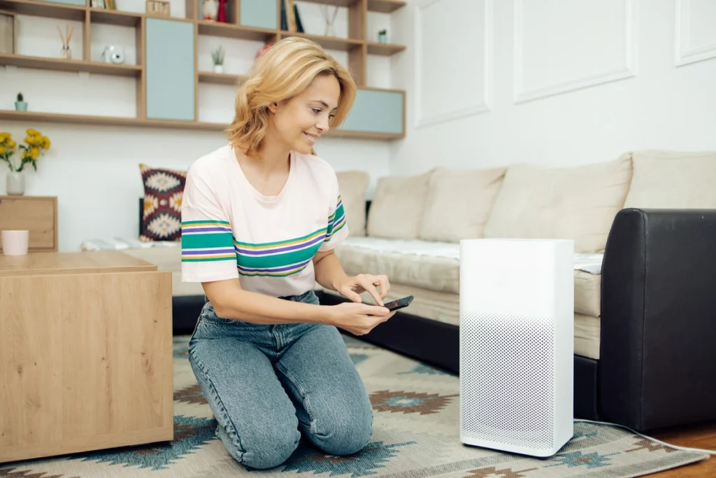 Uśmiechnięta kobieta klęcząca na dywanie w nowoczesnym salonie, sterująca białym, wolnostojącym oczyszczaczem powietrza za pomocą aplikacji w smartfonie.
